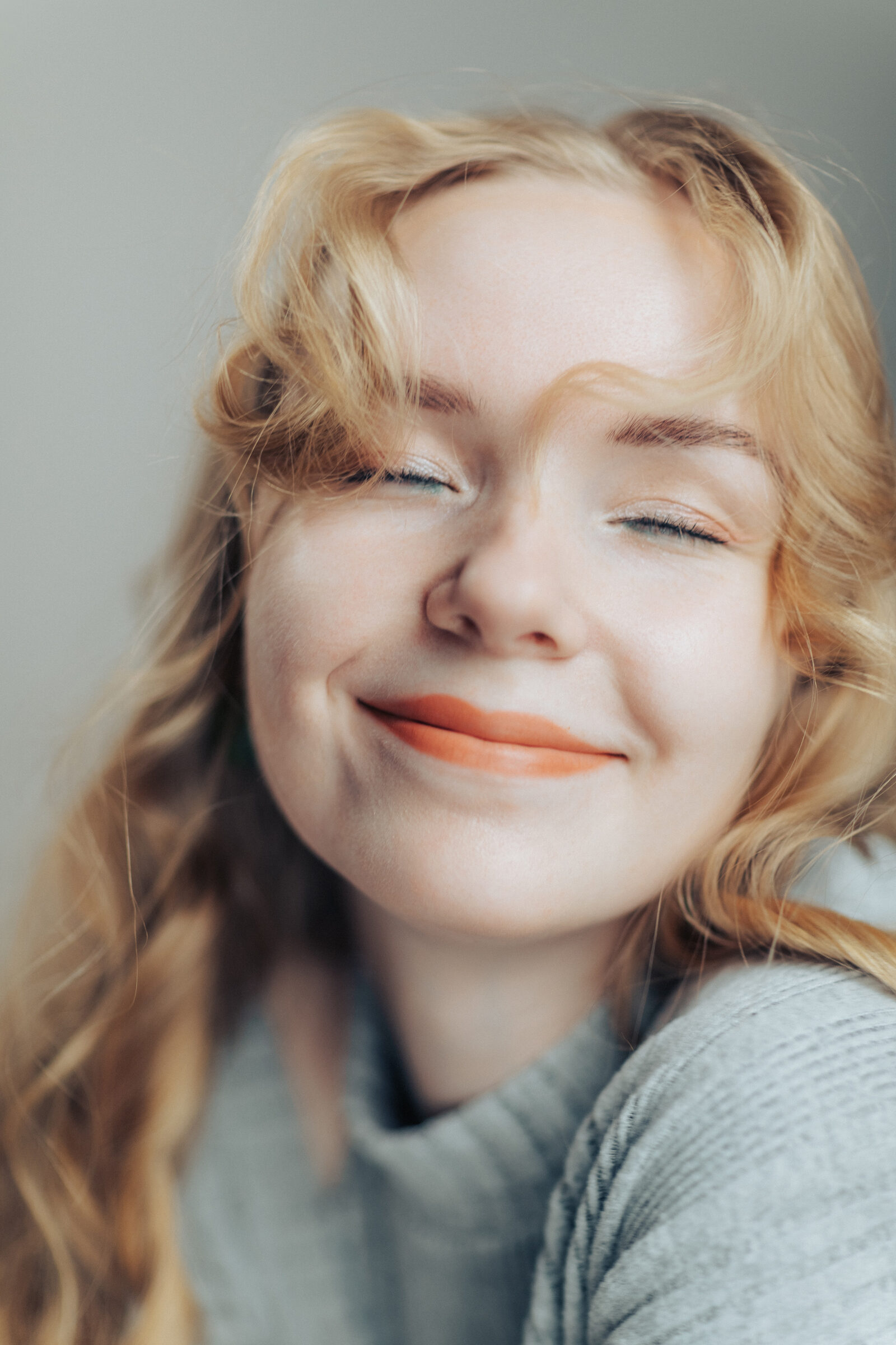 Close portrait of a woman with wavy blonde hair and orange lipstick, eyes closed, smiling